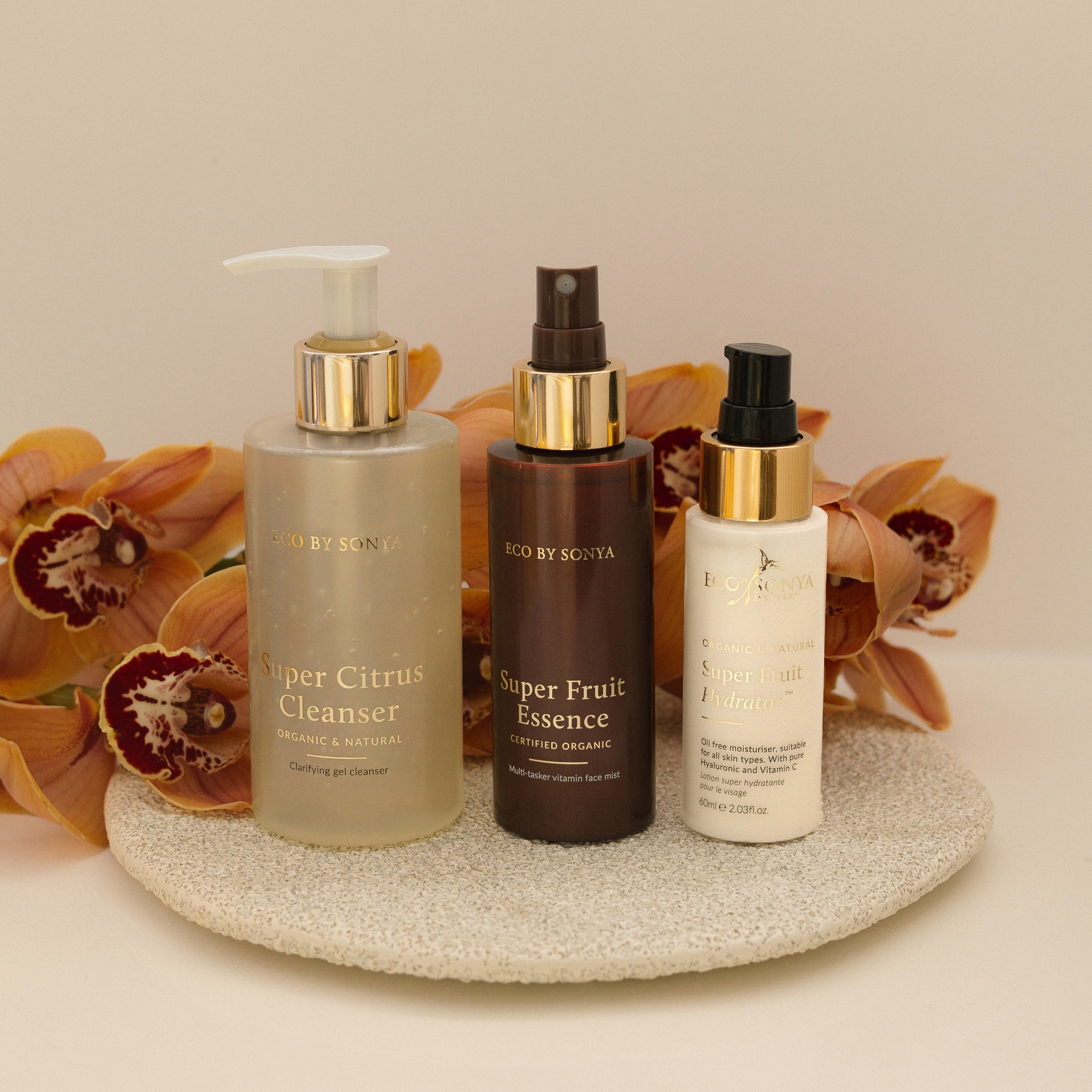 Eco By Sonya 3 step skincare system with Super Citrus Cleanser, Super Fruit Essence and Super Fruit Hydrator on a decorative plate with flowers in the background