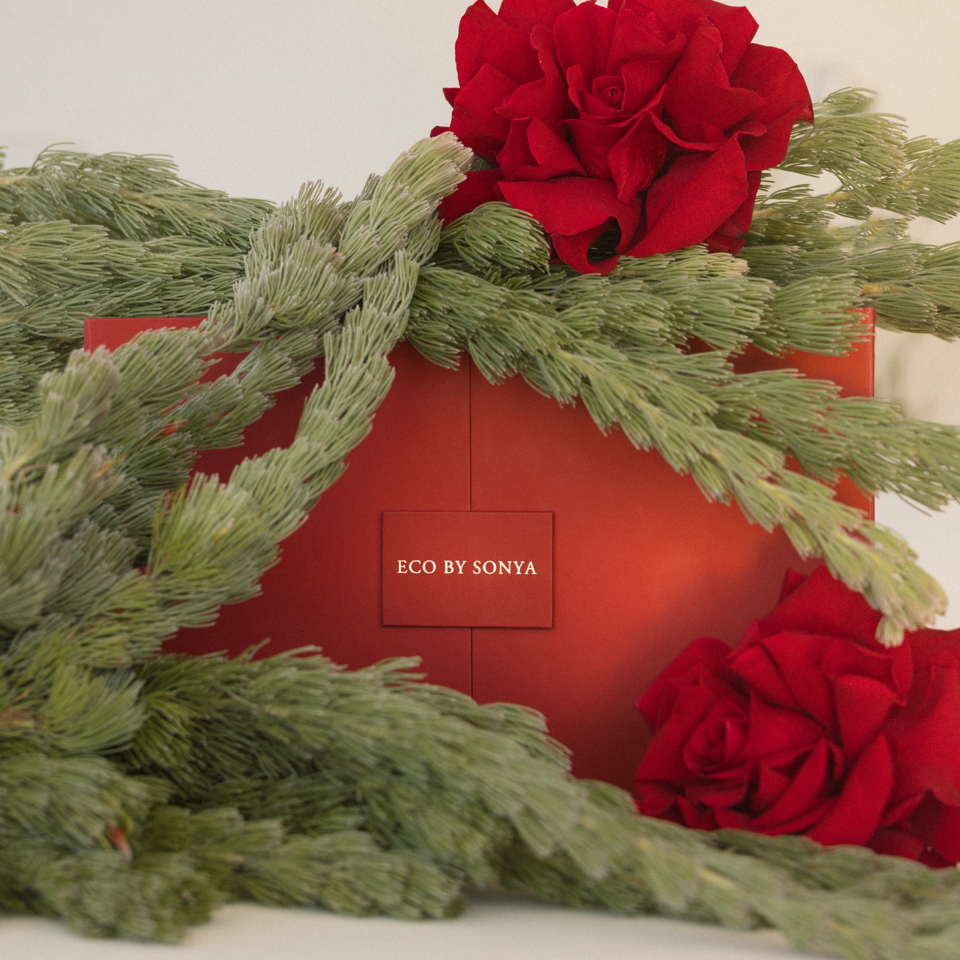 Red advent calendar box with ECO BY SONYA branding, surrounded by red roses and Christmas greenery on a light background.