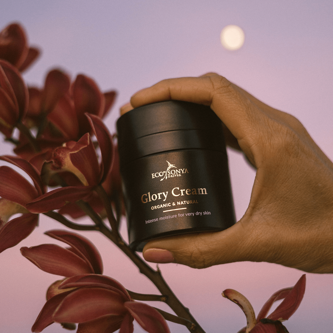 Hand holding Eco By Sonya Glory Cream jar beside burgundy orchid in warm sunset light, natural rich organic face cream.