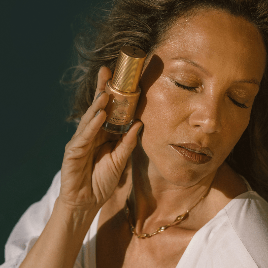 Mature woman with radiant golden skin holding Eco By Sonya Golden Illuminiser bottle near her face.