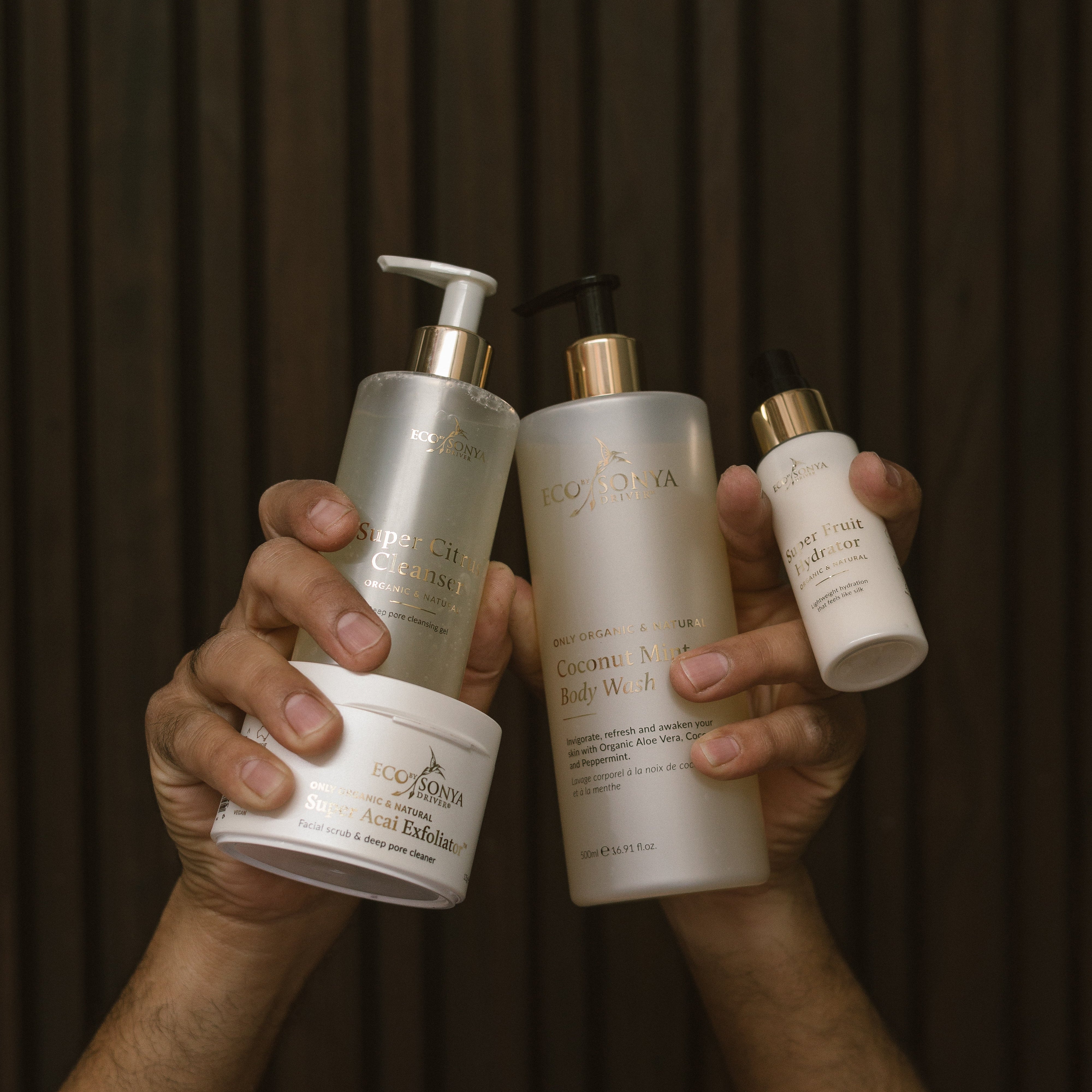 Hands holding various Eco By Sonya skincare products from Young Blokes Pack against a dark background
