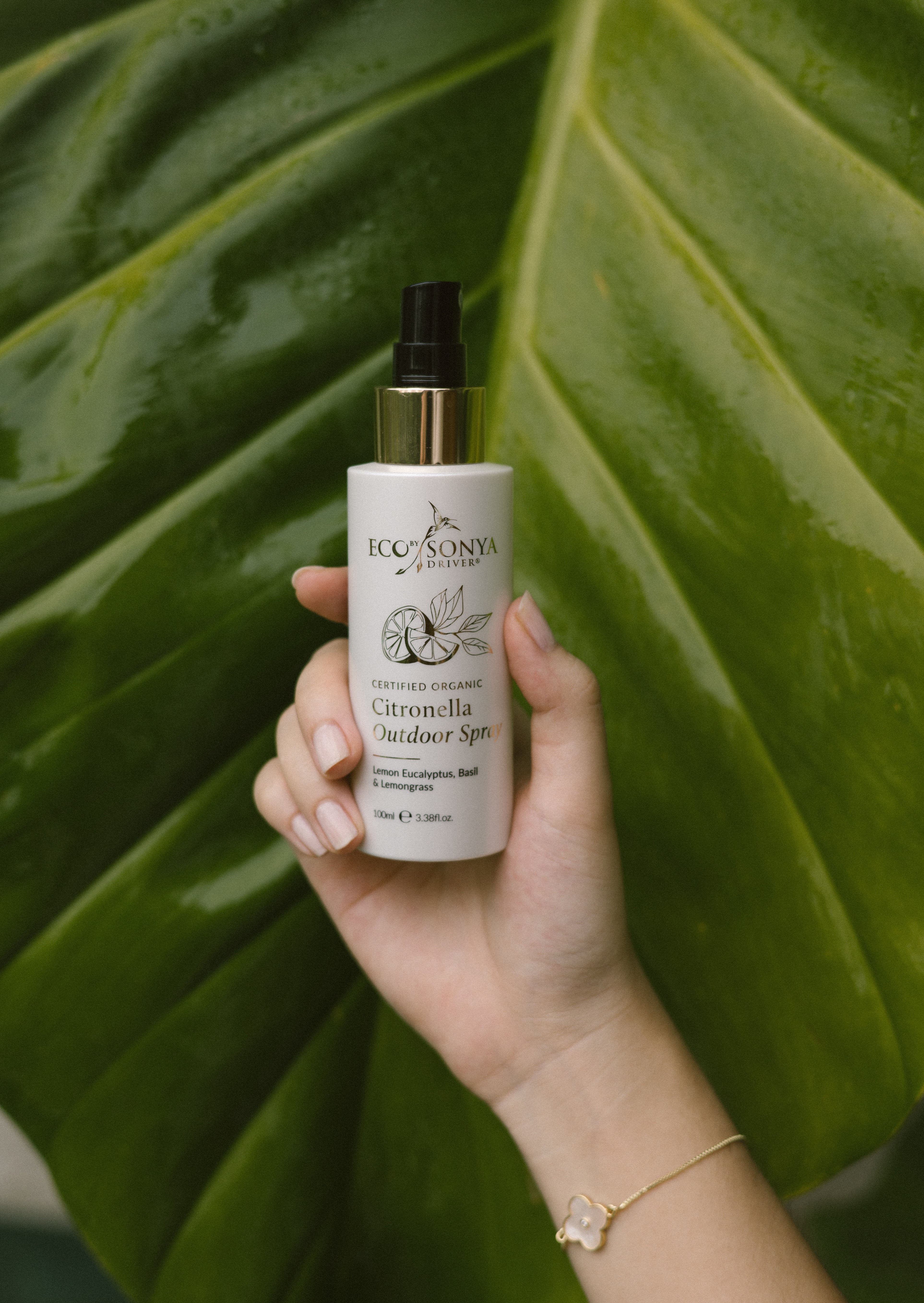 Hand holding a bottle of Eco By Sonya Citronella Outdoor Spray against a green leaf background