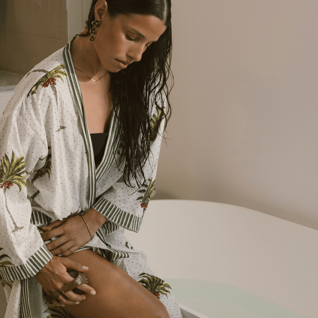 Woman in a patterned robe sitting by a bathtub using body workout massage tool on thigh