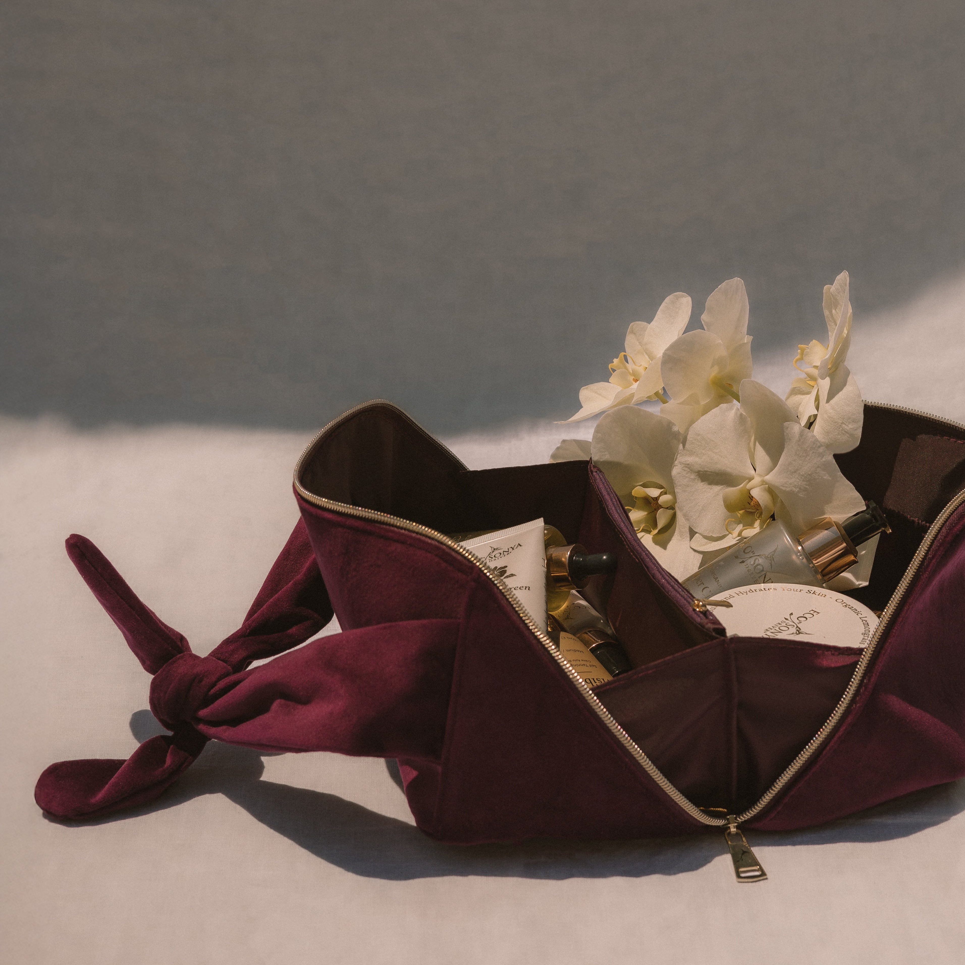 Burgundy travel cosmetic bag open with products inside, white orchids