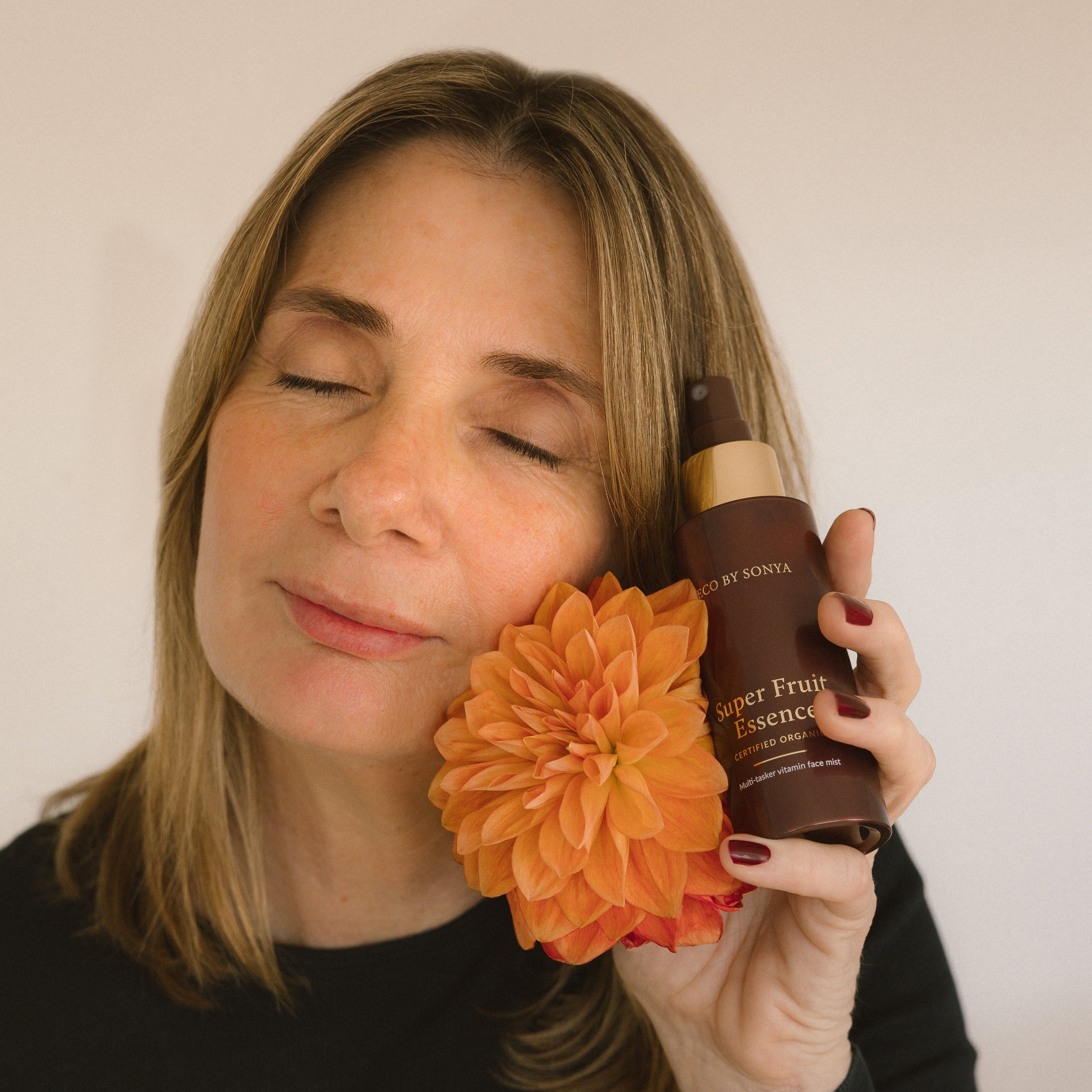 Woman holding an orange flower and a bottle of Eco By Sonya Super Fruit Essence against a plain background