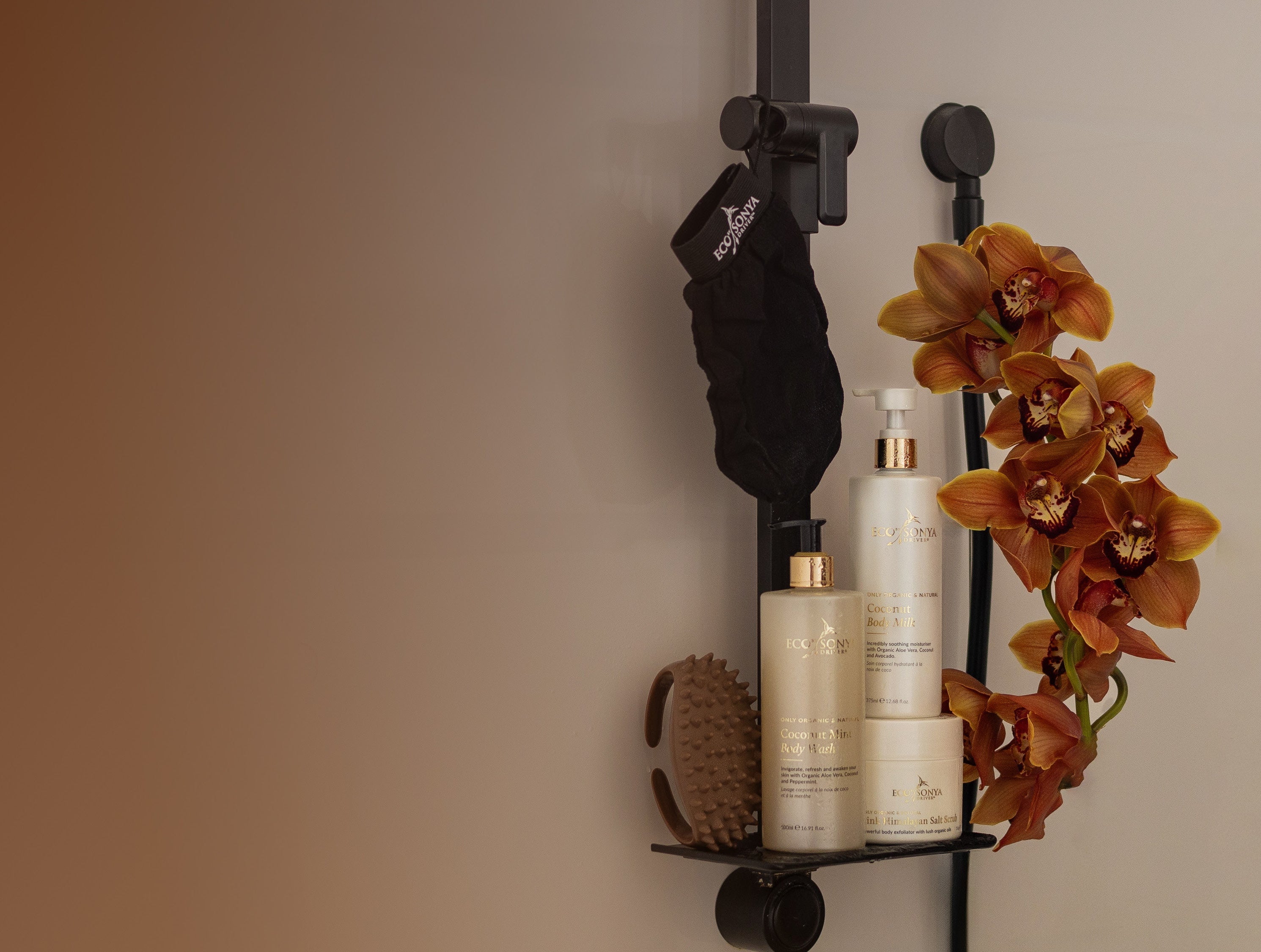 Bathroom shelf with Eco By Sonya Smooth Body Kit products and a flower arrangement against a beige wall.
